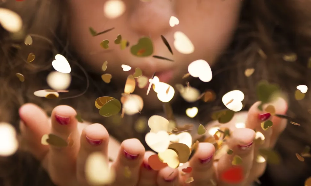 Personne soufflant entre ses mains, sur des confettis dorés en forme de coeur reflétant la lumière.